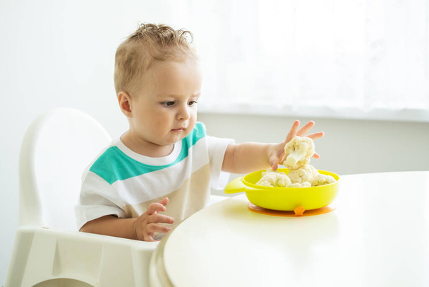 улыбающийся мальчик, сидящий в детском кресле и поедающий цветную капусту на белой кухне - Фото, изображение