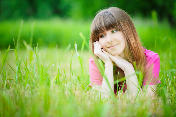 Jeune femme souriante belle dans un chemisier rouge se trouve sur l'herbe sur le fond d'un parc d'été vert. - Photo, image