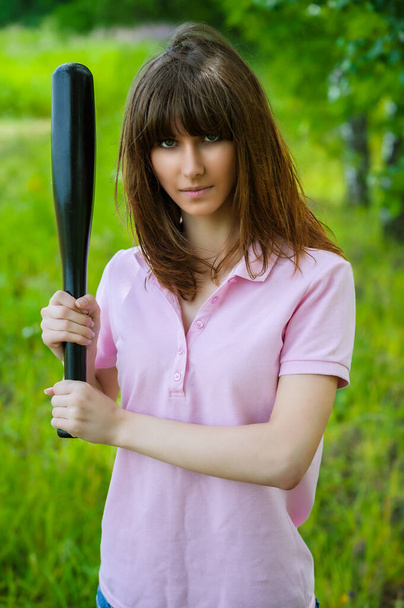 Een mooie jonge vrouw in een blauwe jeans en een roze blouse houdt een zwarte honkbalknuppel tegen een zomergroene achtergrond. - Foto, afbeelding