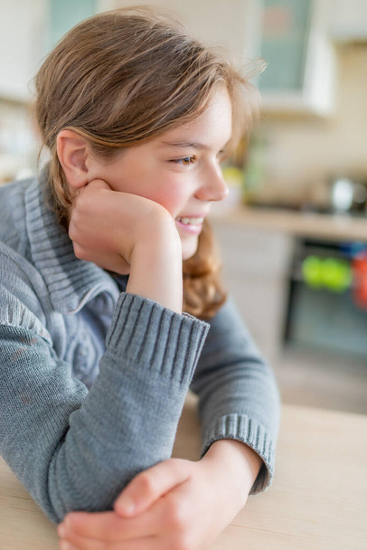Uma linda menina adolescente loira sorridente se senta na cozinha à mesa e olha para um tablet preto. - Foto, Imagem
