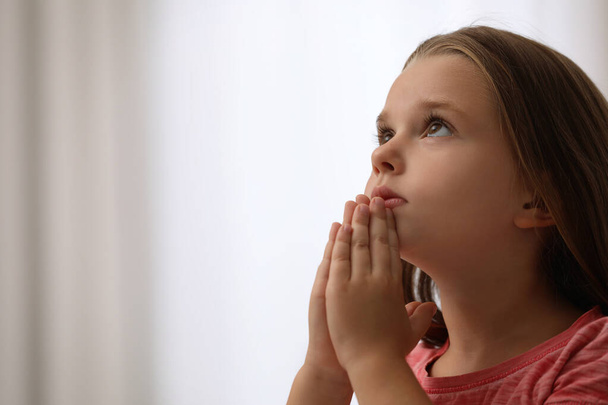 Cute little girl with hands clasped together praying on blurred background. Space for text - Photo, Image