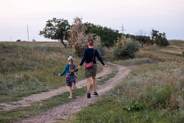 Mother and daughner running back view. Happy family concept. Traveler child with long braided hair in plaid skirt on countryside landscape. Girls walking outdoor on sunset faceless. - Fotografie, Obrázek