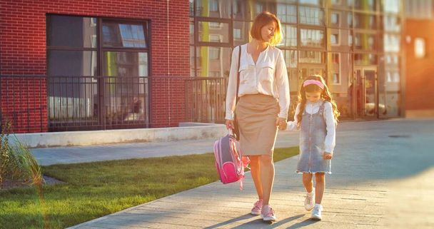 Caucasian mother and cute sad daughter walking from school after lessons outdoor on sunny day. Pretty woman with little upset schoolgirl walk on street in city. Children education concept - Fotó, kép