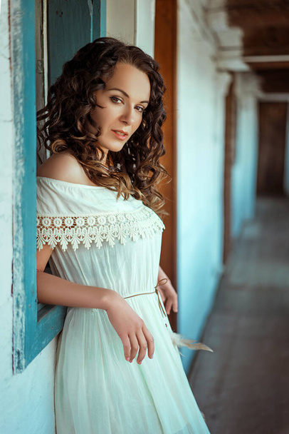 A beautiful Caucasian curly brunette woman wearing a white dress and leaning on the window - Valokuva, kuva
