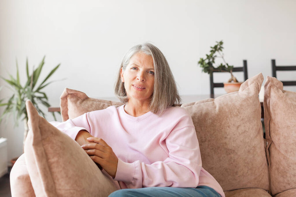Portrait happy healthy middle aged woman sitting on comfortable couch at home. Smiling pleasant 50s elderly gray-haired mother looking at camera, posing in modern living room - Foto, immagini
