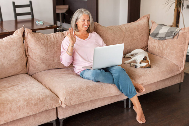 Mature smiling woman waving her hand to laptop while sitting in couch. Female says hello in a video conference chat Happy gray haired middle aged lady at home - Foto, Imagem