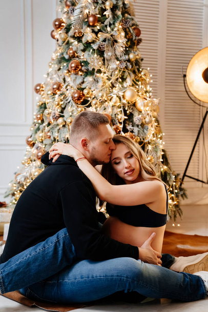 happy man and pregnant woman sitting on the floor by the Christmas tree.  - Fotografie, Obrázek
