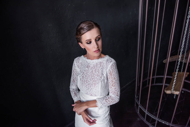 Fashion bride in a white openwork dress without a veil holds a wedding bouquet in her hands. Young woman stands by a large iron birdcage on a black textured background. Fictitious marriage. Copy space - Foto, Imagen
