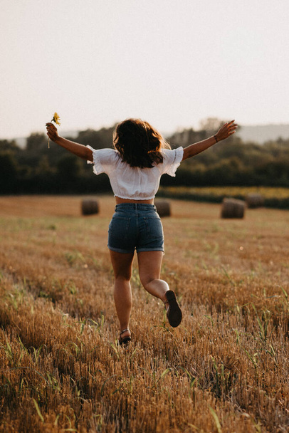 A young brunette girl with shorts and a white top happily running in a wheat field during sunset - Fotó, kép