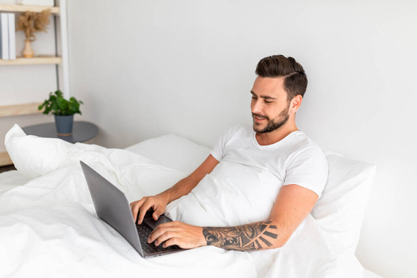 Δουλειά στην κρεβατοκάμαρα, σέρφινγκ, περιήγηση στο διαδίκτυο. Ευτυχισμένος χιλιετής άνθρωπος ξύπνησε, κάθεται στο λευκό κρεβάτι, ελεύθερος χώρος - Φωτογραφία, εικόνα