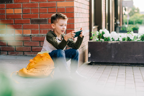 Wracać do szkoły. Słodkie dziecko z plecakiem, trzymające telefon komórkowy, bawiące się telefonem komórkowym. Uczeń z torbą. Uczeń podstawówki po zajęciach. Dzieciak siedzi na schodach na zewnątrz na ulicy - Zdjęcie, obraz