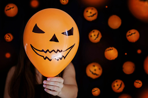 Automne, saison et concept d'Halloween - femme tenant un ballon orange et couvrant son visage sur fond noir avec des lanternes d'Halloween orange - Photo, image