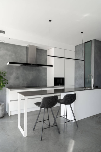 Spacious kitchen with minimalist square shaped furniture, grey floor and walls, counter with integrated sink and pair of bar stools in front of it, built-in microwave and cooktop. Vertical shot - Foto, immagini