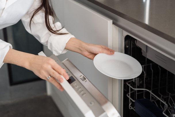Καλλιεργημένη φωτογραφία της γυναίκας που χρησιμοποιεί σύγχρονο ενσωματωμένο πλυντήριο πιάτων, ενώ κάνει οικιακές εργασίες στην κουζίνα στο σπίτι, βάζοντας λευκό πιάτο μέσα - Φωτογραφία, εικόνα