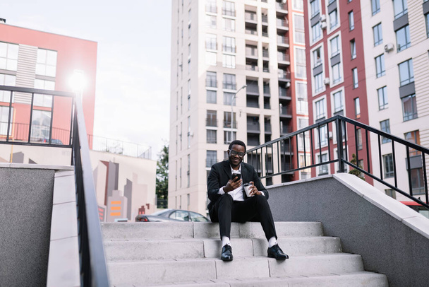 Ένας νεαρός άνδρας στα σκαλοπάτια ενός κτιρίου κάθεται με ένα τηλέφωνο, κρατάει καφέ, επικοινωνεί με φίλους και εργάζεται στο δίκτυο - Φωτογραφία, εικόνα
