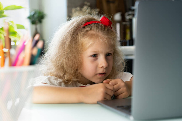 μικρό κορίτσι κάνει μαθήματα στο σπίτι σε ένα φορητό υπολογιστή, γράφει με ένα στυλό σε ένα σημειωματάριο. - Φωτογραφία, εικόνα
