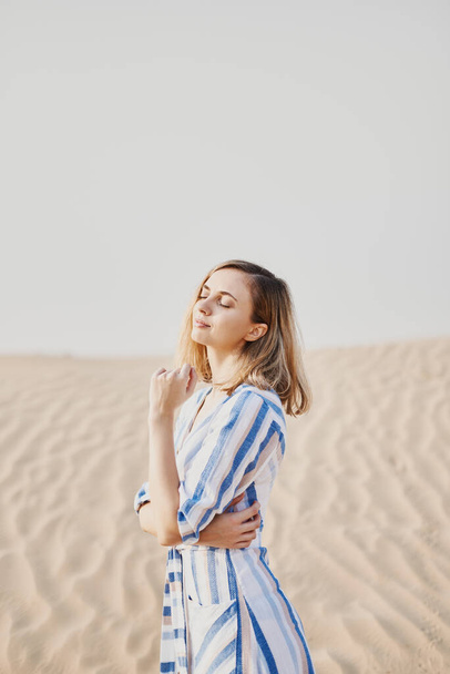 Portrait of a young woman in a blue dress standing alone in a sandy desert and enjoying sun, weather and wild desert atmosphere - 写真・画像