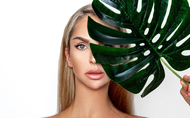 A beautiful young girl in the studio on a white background with flawless and healthy skin holds a large green tropical leaf in hands and covers a part of her face. Fashion, beauty, make-up, cosmetics. - Fotografie, Obrázek
