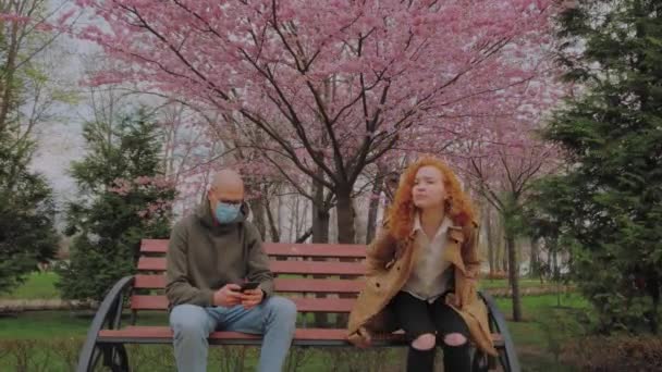 European Man with mask and woman without mask sitting on bench in park. Woman coughs contagious. Coronavirus epidemic - Footage, Video