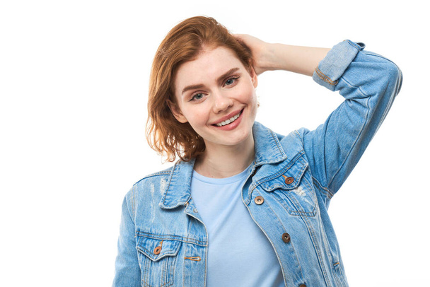 A girl student with red hair happily thinks and looks at the camera while holding her finger near her chin. Pensive thoughtful woman in a blue denim jacket on a white background. Copy space - Foto, afbeelding