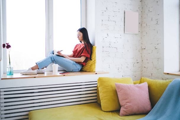 Thoughtful female in glasses wearing casual clothes taking notes in notebook while sitting on windowsill next to flowers in modern apartment - Foto, Bild