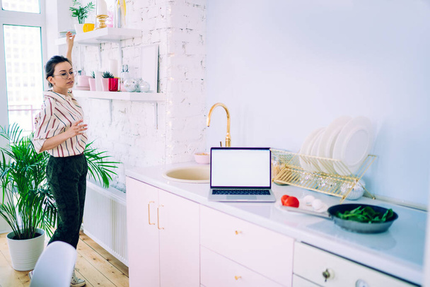 Side view of young female in casual outfit taking mugs from shelves and looking away while standing near counters with salad ingredients and laptop with blank screen in kitchen - Fotó, kép