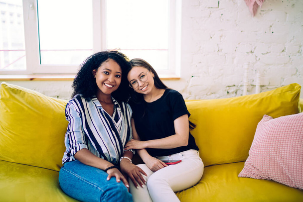 Multiethnic female teenagers smiling and looking at camera while sitting together on comfortable couch near window in stylish modern apartment - Photo, Image