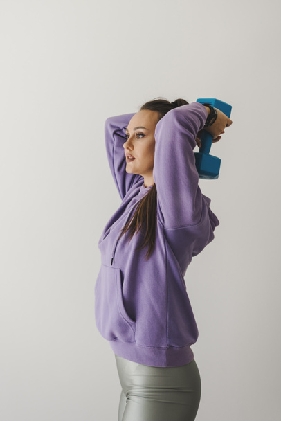 Young female in purple hoodie and green shorts exercising with a dumbbell. Indoor workout with equipment - Photo, Image
