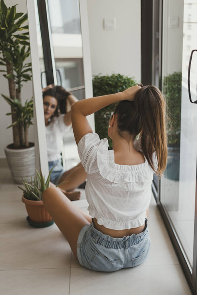 Young woman with long hair sitting on the floor in front of the mirror and making a pony tail. Morning routing, self-care, mindfulness concept  - Photo, image