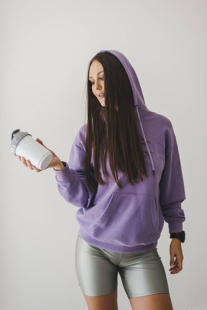 Spor kıyafetli genç ve aktif Avrupalı bir kadın elinde plastik bir su şişesi ya da protein karıştırıcı tutuyor. Egzersiz öncesi veya sonrası proteini - Fotoğraf, Görsel
