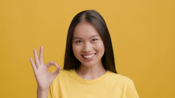Alegre asiático hembra gestos bien sonriendo a cámara, fondo amarillo - Metraje, vídeo