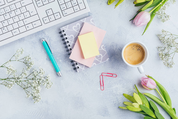 Arbeitsplatz mit Tasse Kaffee, leerem Notizbuch, Tastatur, Zubehör und Blumen in hellem Ton - Foto, Bild