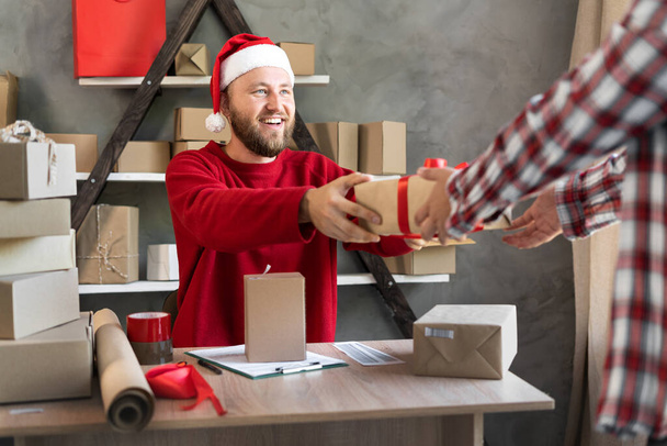 Beginnen met het MKB-concept. Jonge mannelijke aspirant-ondernemer, kleine ondernemer, geeft een doos aan een klant uit een magazijn. Gekleed in een kerstmuts te koop in het kerstseizoen. kopieerruimte. - Foto, afbeelding