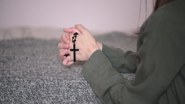 Жінка брюнетка молиться тримає чорний розарій з хрестом в руках і цілує символ, що стоїть на колінах сірим диваном крупним планом
. - Кадри, відео