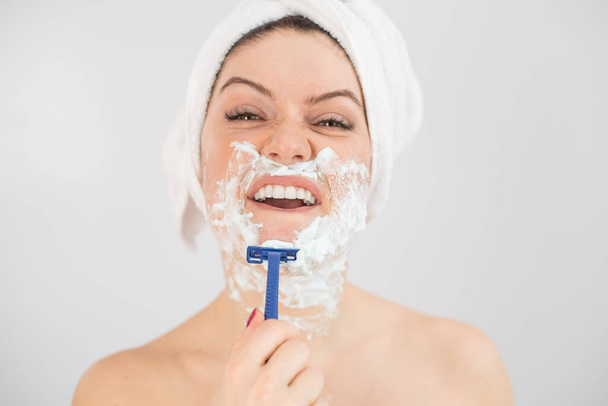 A woman with a white terry towel on her head and with shaving foam on her face shaves on a white background - Fotó, kép