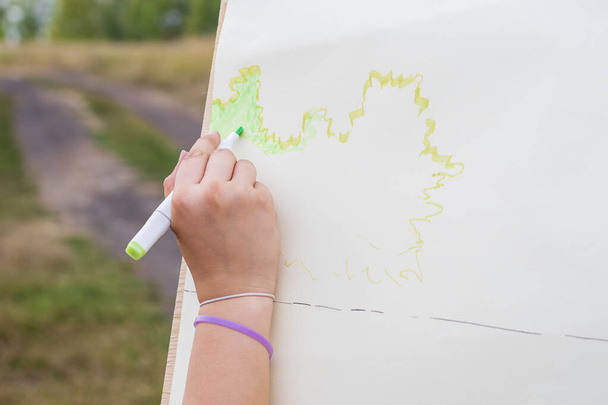 A woman's hand draws a sketch of the future drawing with a green marker on the white surface of the paper. In the background there is a dirt road and foliage of trees. - Photo, Image