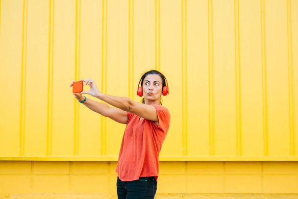 Femme d'âge moyen portant des écouteurs rouges et des vêtements rouges prenant un selfie avec un smartphone rouge sur un fond jaune - Photo, image