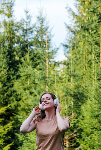 Young woman enjoys listening to music with headphones in forest in nature. Portrait young beautiful caucasian woman dreaming listening to music via headphones with closed eyes outdoor - Photo, image