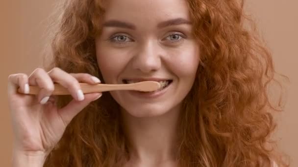 Mujer pelirroja con tirantes que limpian los dientes con cepillo de dientes, fondo beige - Imágenes, Vídeo