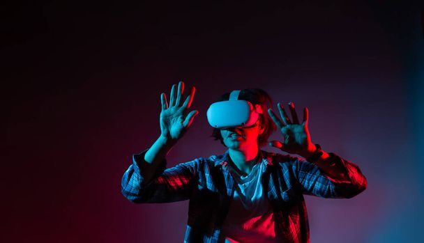 Белая женщина 35-40 лет в очках, используя гарнитуру виртуальной реальности, пытается прикоснуться к чему-то рукой в неоновом свете. - Фото, изображение