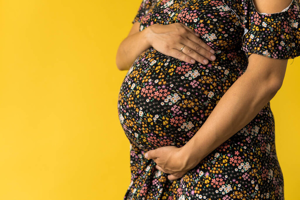 Moederschap, vrouwelijkheid, liefde, zorg, wachten, hete zomer - helder bijgesneden Close-up onherkenbare zwangere vrouw in bloemen zwarte jurk met handen over buik wrijven buik op gele achtergrond, kopieer ruimte - Foto, afbeelding