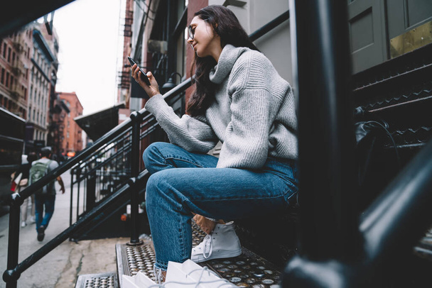 Side view of young woman in gray sweater and blue jeans and eyeglasses sitting on stair while surfing mobile on street of New York city  - Fotó, kép