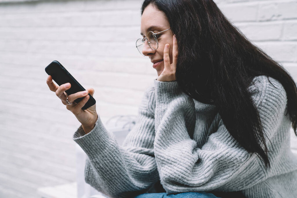 Crop young cheerful woman in grey pullover and glasses sitting next to shopping bags on bench smiling and chatting on cellphone outdoors in New York - 写真・画像