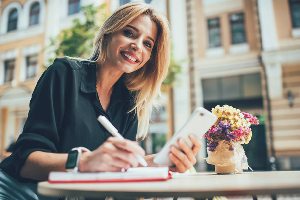 Portrait of cheerful female planning organisation in sidewalk cafe using smartphone device for information research,prosperous woman with cute smile holding cellphone and posing on leisure - 写真・画像