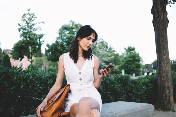 Pensive brunette woman in white dress and elegant bag surfing mobile phone sitting on bench in street with green fence  - Photo, Image