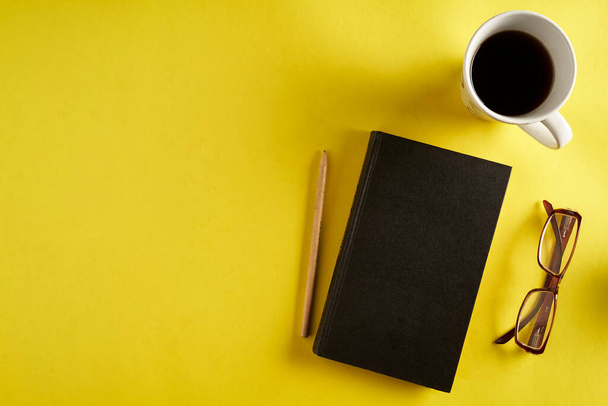 top view of yellow surface, desk with pencil, black notebook and cup of coffee - Photo, Image