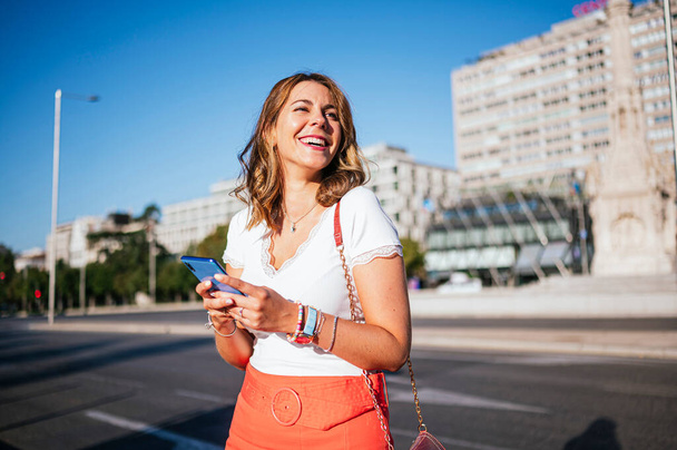 Seitenansicht einer Frau in Sommerkleidung, die ihr Smartphone benutzt und in der Stadt steht. - Foto, Bild