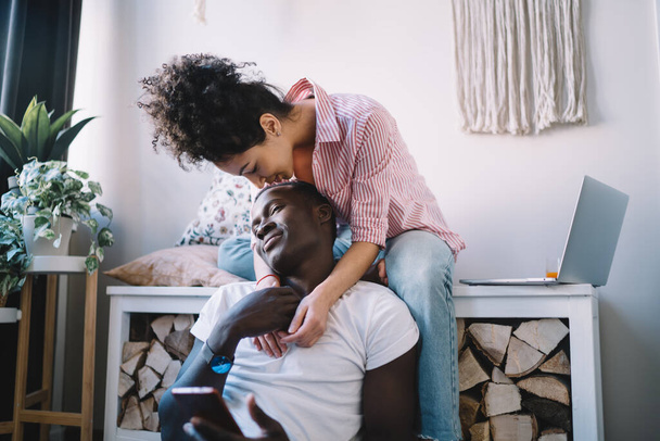 Young curly woman with bun wearing striped shirt hugging from back and kissing on head relaxed smiling black man sitting with smartphone in hand - Photo, Image
