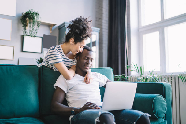 Smiling ethnic female hugging black boyfriend with tender while spending time at home and using modern laptop together in weekend - Foto, Imagen