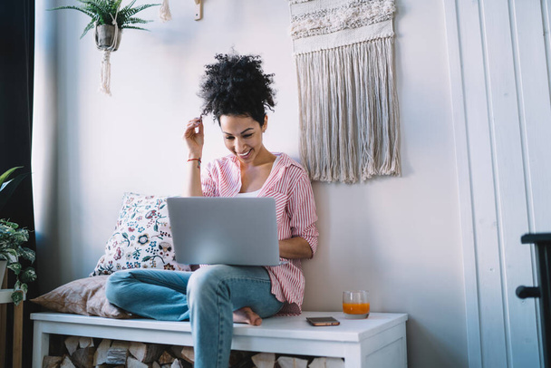 Laughing focused ethnic female in casual clothes having rest on trendy couch and using laptop in modern flat while enjoying comedy - Foto, afbeelding